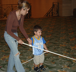 Inspired by the cane travel teachers she worked with at convention, Melissa Fernandez teaches her son Jacob about using the long white cane. He is pictured here astride the cane like a horse. Perhaps he has a little more to learn about cane use.