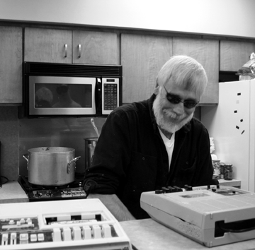 Pete Hoffman smiles at the camera while he works. Behind him can be seen his pots of goulash on the stove.