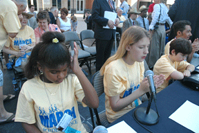 Marché Daughtry, Hannah Weatherd, Jordan Richardson, and Vejas Vasiliauskas are seated at the head table on the rally stage. They presented a resolution on the importance of Braille and the NFB as tools to help their generation gain a quality education and solid employment. Hannah Weatherd is speaking into the microphone while the others listen.