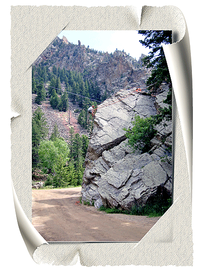 In the foreground we can see a large rock formation and a man scaling the rock to anchor the fixed ropes the climbers will use. In the background are tree-covered mountains. Below the rock winds a dirt path that gives access to the park.
