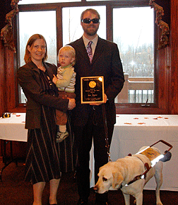 Amber and Dan Bigley stand together at the awards banquet. She is holding their son while Dan displays his award plaque.