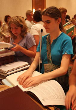 A teenager is reading a Braille book that is lying open on a stack of books. A number of people can be seen around her.