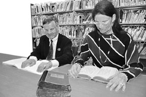 A man reading Braille and a woman reading print are seated at a table in the Jacobus tenBroek Library with bookshelves filled with volumes behind them and a stack of Braille books on the table in front of them.