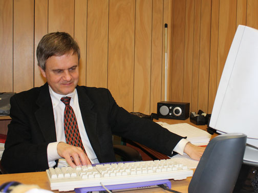 Steven Booth grades a hard-copy Braille certification lesson and writes his report using the refreshable Braille display and JAWS on his computer.