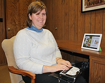 Amy holds a Braille notetaker in her lap while she types into and reads from an iPad on her desk.
