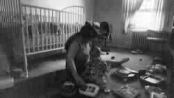 Photo of a mother and baby playing in the nursery with many toys and crib in the 
background.