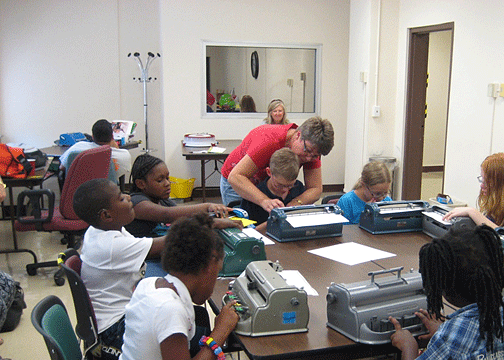 Nine students sit around a large table; each student is writing on a Braille writer. A teacher helps one student.
