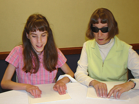 A blind teen and a blind adult sit at a table reading a Braille passage together.