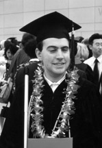 Photo of Mark Riccobono standing in his cap and gown holding his diploma.