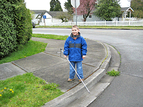 Since an important aspect of the cane is to detect drop-offs, utilizing a longer cane is beneficial. Here a five-year-old boy proudly displays his long white cane as he locates the curb.