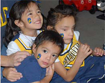 Milagro sits on the floor with her little sister Kotomi and little brother Kiyoshi. All of the children have their faces painted, and the girls wear cheerleading uniforms.