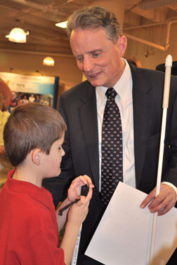 Holding a sheaf of Braille pages, President Maurer talks with a boy in a red shirt in the exhibit hall. Both are holding long NFB canes.