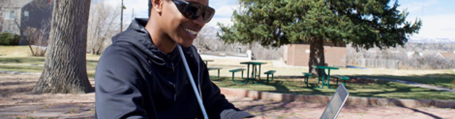 A young blind man sits at a picnic table in the park using a laptop.