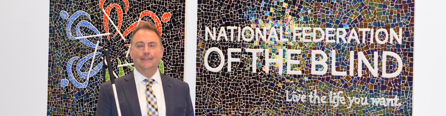 Mark Riccobono stands in front of a mosaic of the National Federation of the Blind logo and tagline live the life you want.