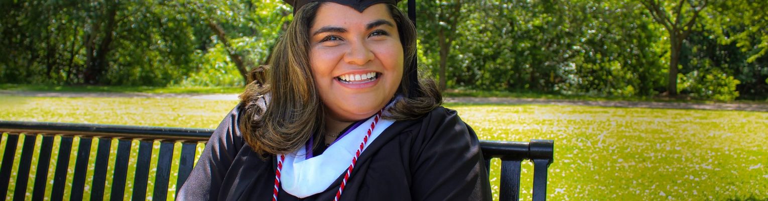 A smiling Kenia Flores in her graduation cap and gown sits on a bench outside.