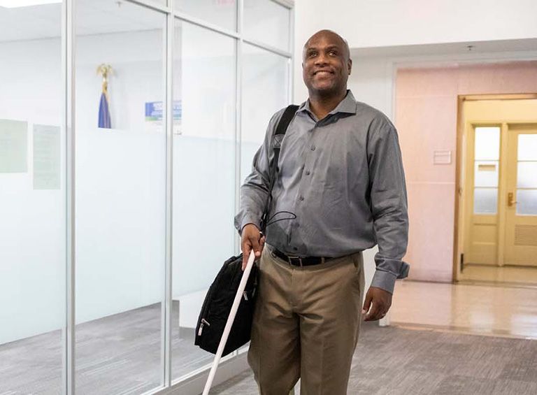 Blind man in business attire walks with white cane and brief case in office hallway
