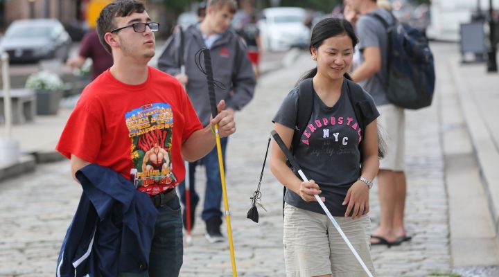 Two blind people walk on a sidewalk using white canes.