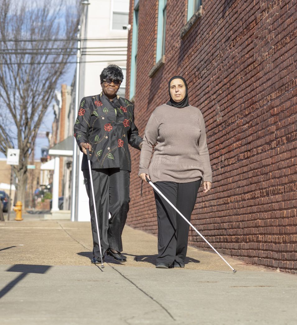 Two women use their white canes to walk down the street.