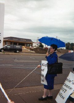 Joyce Scanlan, wearing a picket sign hanging from her neck, walks with a cane and carries an umbrella.