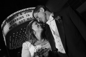 Couple, still in their wedding dress, nuzzle noses outside a movie theater.