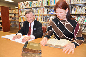 A man reading Braille and a woman reading print are seated at a table in the Jacobus tenBroek Library with bookshelves filled with volumes behind them and a stack of Braille books on the table in front of them.