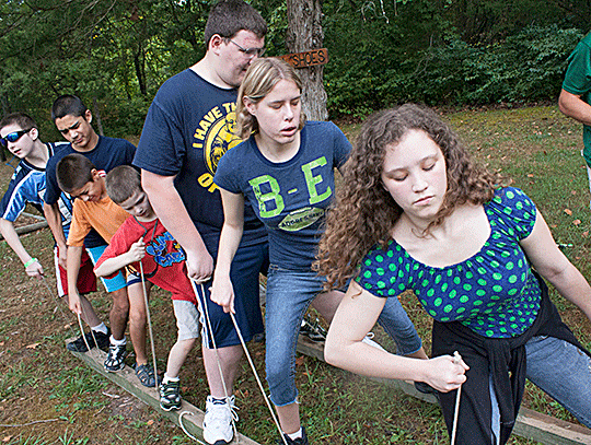 Seven young people stand  in a line, left feet on a two-by-four plank, right feet on a parallel two-by-four  plank, each side controlled independently by ropes held in the students&rsquo; hands.  The object is to walk forward by picking up each side in turn, which requires  concentration and much cooperation.
