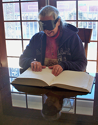 Kate Smith sits in the dining room of the Louisiana Center for the Blind reading a Braille book while wearing the sleepshades used in training.