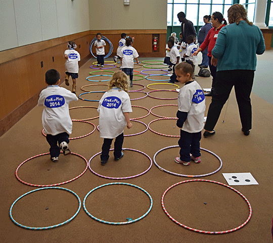 There are hula hoops on the ground in patterns that represent the shapes of Braille cells, and the Pre-K children are standing in the hoops to match the print letters shown on the papers inside them.
