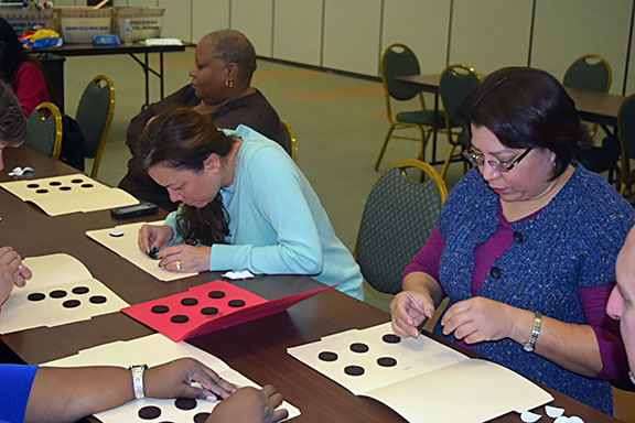 Ellen Bedford, Kathryn Webster, and Joy Scott participate in one of the &ldquo;Taste of NFB BELL&rdquo; sessions, playing Braille Twister. People-sized tactile Braille cells were created on the floor using Poly Spots (that&rsquo;s the official term for these little rubber discs that PE teachers usually use to mark bases or spots on the floor for various games). In this game, participants use parts of their body, like in Twister, to mark dots to create Braille letters, contractions, or other symbols. In this picture they are using their feet to make the letter &ldquo;b.&rdquo;
