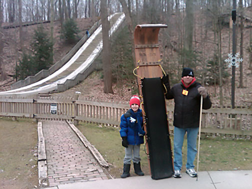 David and Matthew pose in front of the toboggan shoots. They are holding the big, long toboggan between the two of them.