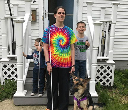 Heather Bird stands in front of her house with her long white cane and her guide dog. Her two young sons, one on each side, hold their own child-sized canes.