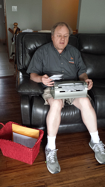 Gary Wunder is shown with a Braillewriter on his lap, a piece of mail in his right hand, and a mail basket at his feet. 