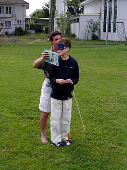 Standing behind a boy wearing sleepshades, Daniel holds a hardcover picture book a few inches from the boy&rsquo;s right side.&nbsp;The student has turned his head in response.&nbsp;