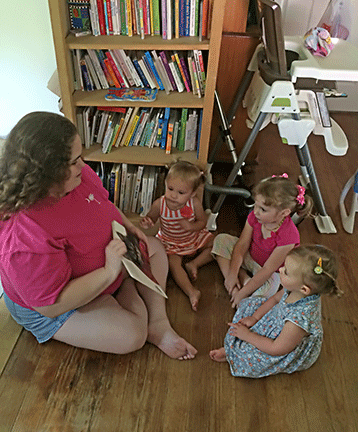 Sitting on the floor, Brandy reads a picture book to three little girls gathered around her.