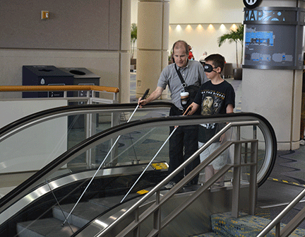 A mobility instructor teaches a young blind teen to use an escalator.