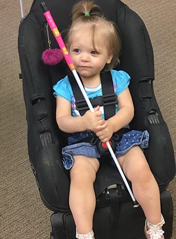 A small child sits in a car seat, holding her cane.