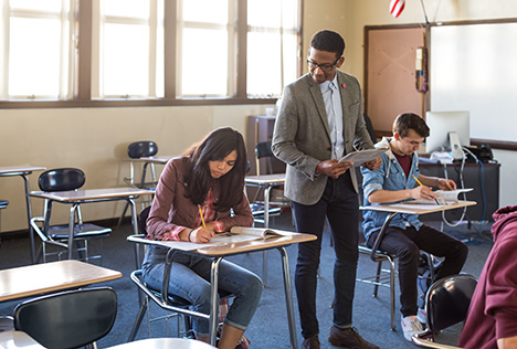 Two students take a standardized test while a teacher looks on.