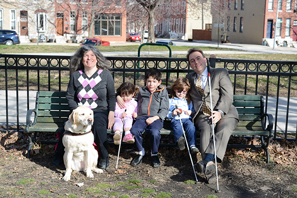 Mark and Melissa Riccobono sit on a park bench with their three children. Melissa&rsquo;s guide dog is beside her.